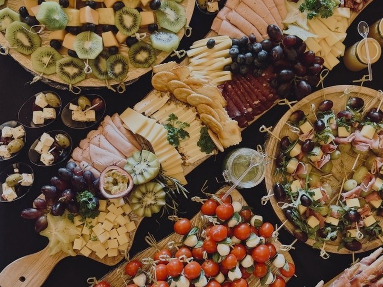 Draufsicht auf reichhaltig gedeckte Apéro-Platten mit Fruchtspiessen, Tomate-Mozzarella-Spiessen, Aufschnitt, Grissini und Trauben auf schwarzem Grund