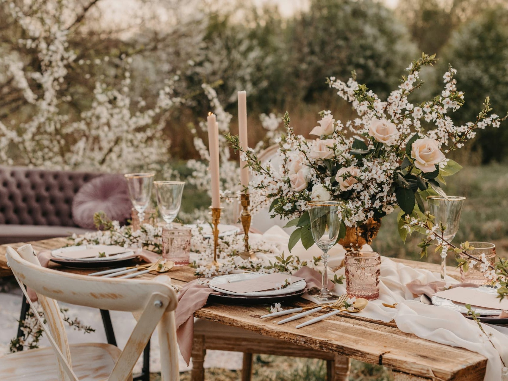 Holztisch im Garten mit Kirschblüten, weiss-rosafarbenem Blumengesteck in goldener Vase, hellrosa Kerzen in goldenen Leuchtern und gedeckten Tellern