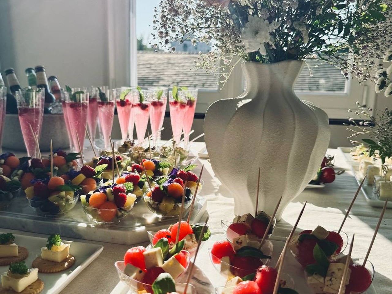 Sektflöten mit rosafarbenem Drink neben Häppchenschälchen mit Melone, Feta und Minze, dahinter eine Vase mit Sommerblumen