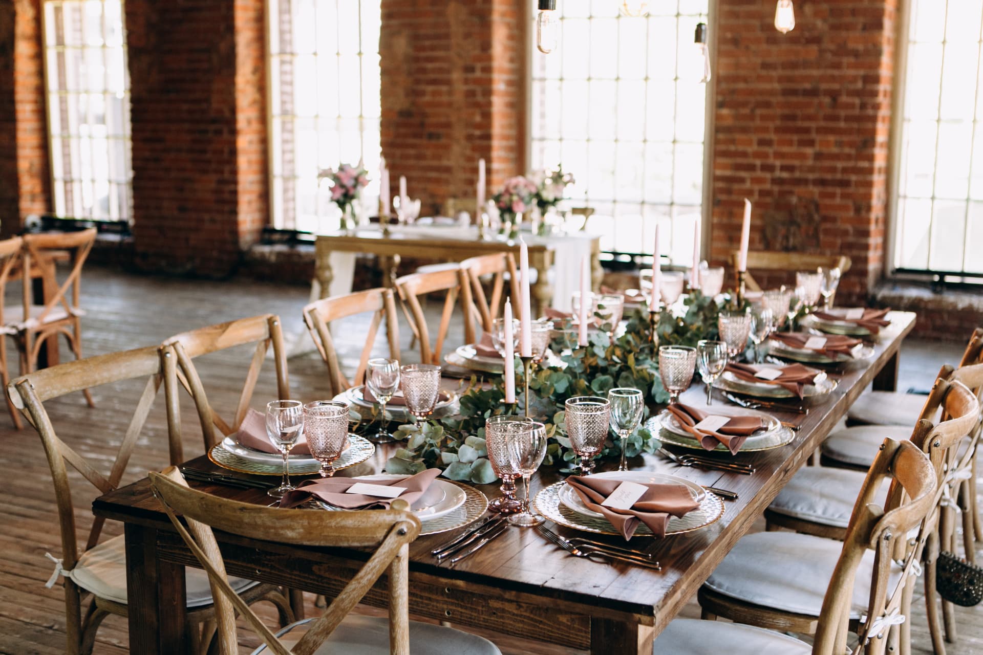 Gedeckte Festtafel mit Eukalyptus-Läufer, rosafarbenen Kerzen in goldenen Leuchtern, rosafarbenem Glas und Holzstühlen in einem Loft mit Backsteinwänden und grossen Fenstern