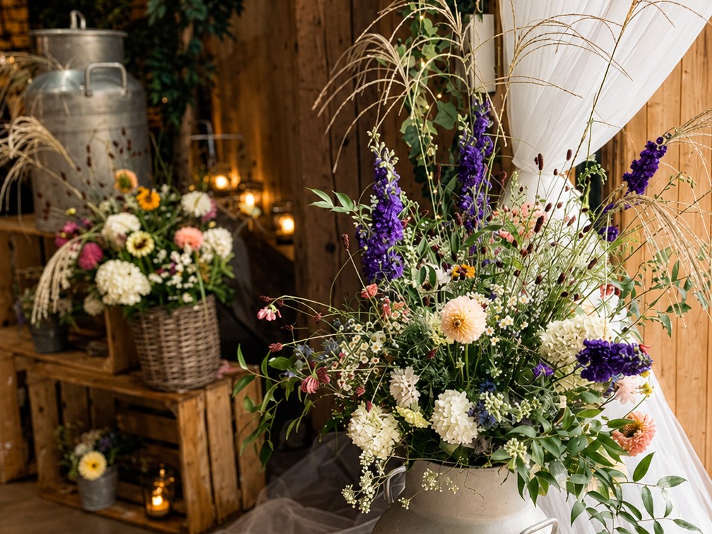 Milchtrenne mit hohem Blumengesteck aus violettem Rittersporn vor weissem Tüll und Holzkisten in einer Scheune
