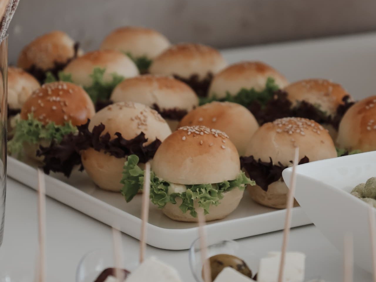Mini-Sliders mit Sesam auf einer weissen Platte, davor Becher mit Feta und Oliven am Spiess