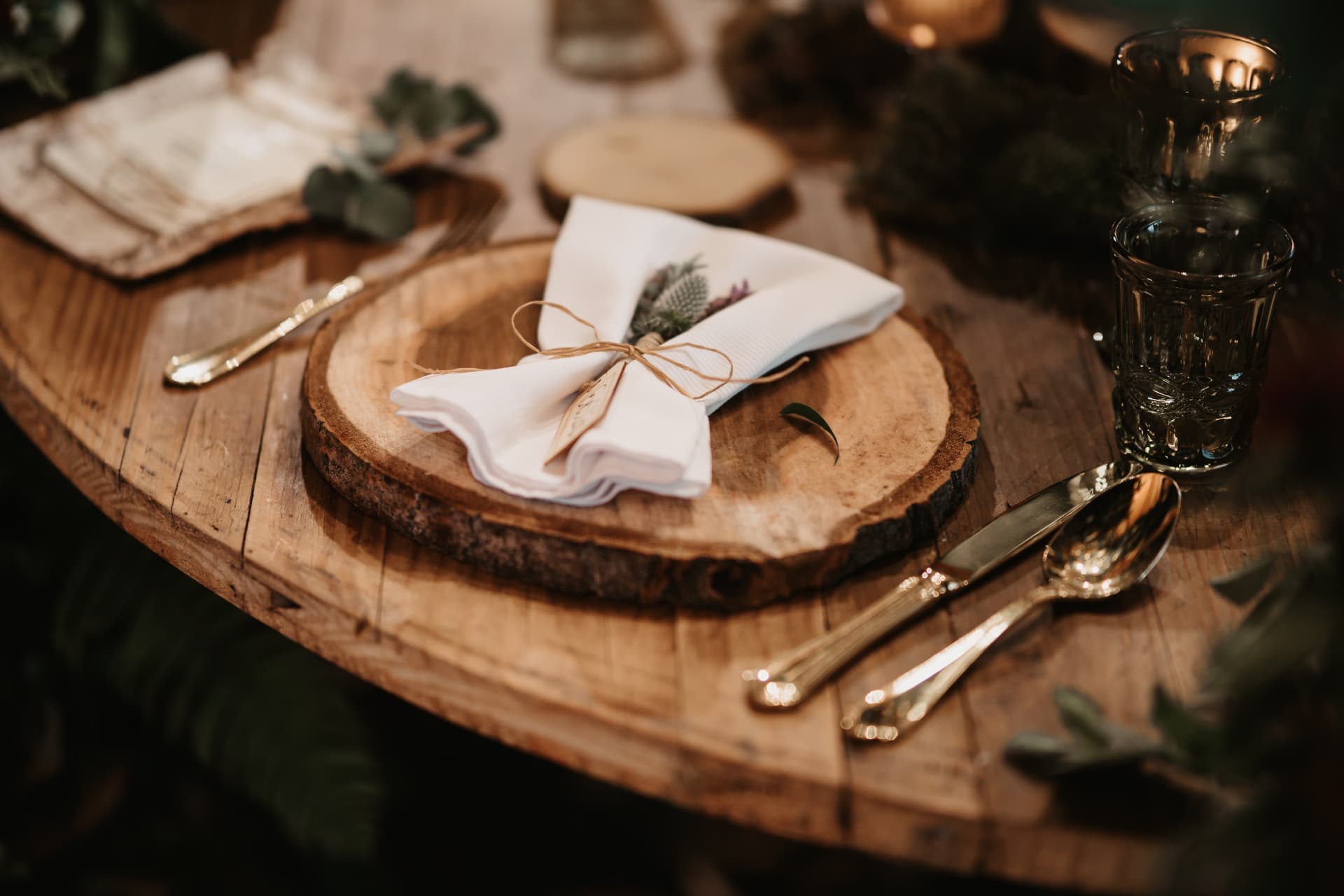 Gedeckter Holztisch mit Baumscheibe, weisser Serviette mit Namenskärtchen und Trockenblume sowie goldfarbenem Besteck