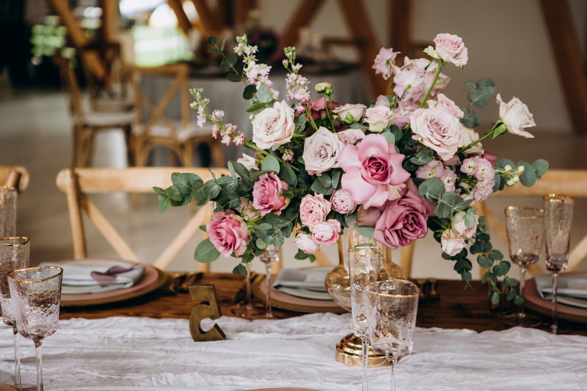 Festlich gedeckter Holztisch in einer hellen Scheune mit Blumengesteck aus altrosafarbenen Rosen und Eukalyptus, Gläsern mit Goldrand und Tischnummer