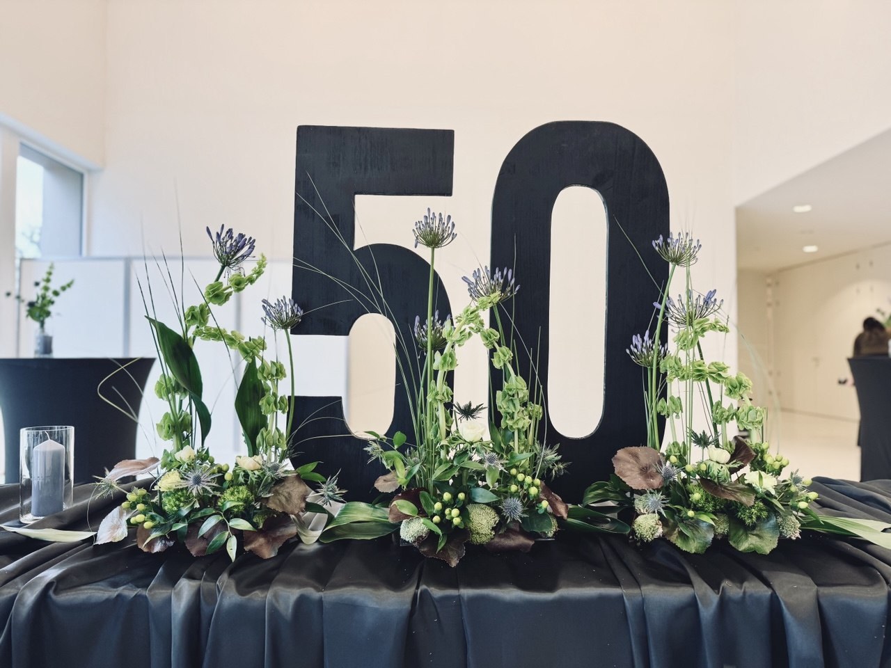Schwarze Holz-Ziffern 50 vor drei Blumengestecken auf schwarz gedecktem Tisch