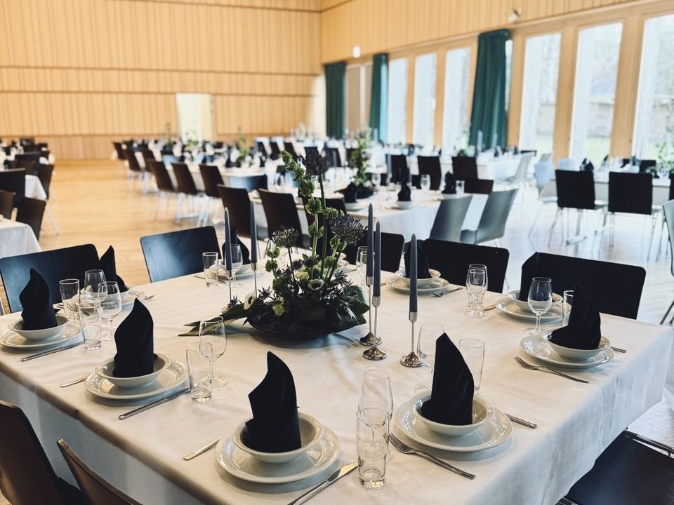 Festlich gedeckter Festsaal mit weiss eingedeckten Tischen, aufrecht gefalteten schwarzen Servietten, dunklen Stabkerzen und Blumengestecken mit Disteln, dahinter weitere Tische und eine hohe Fensterfront