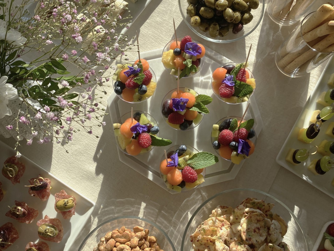 Draufsicht auf ein Apéro-Buffet mit Marmorteller voller Fruchtbecher und essbaren Blüten, umgeben von Crackern, Oliven, Grissini und Nüssen