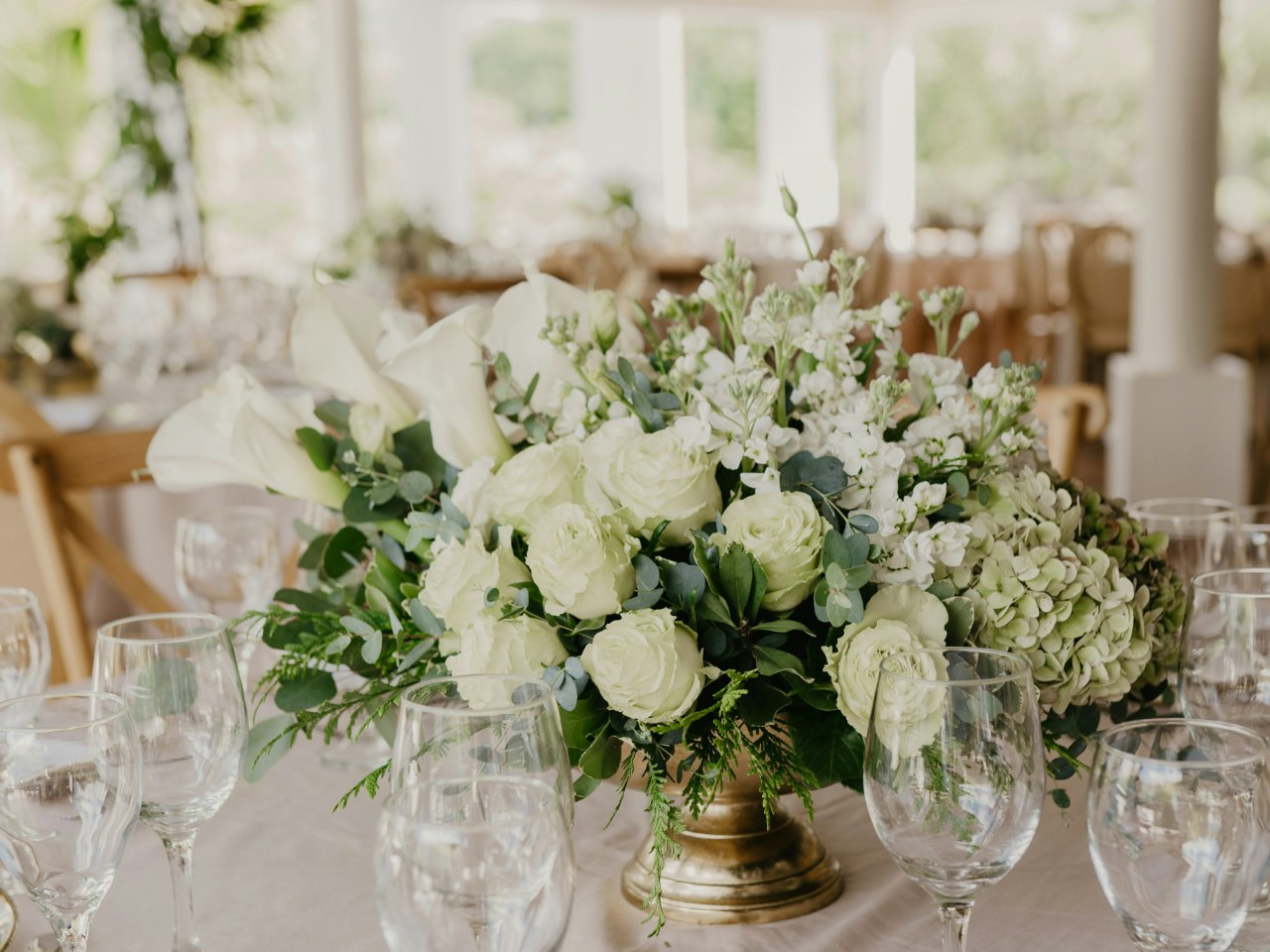 Blumengesteck mit weissen Rosen, Calla und Hydrangea in goldener Vase auf rosa gedeckter Tafel mit Menükarten auf Goldrandtellern