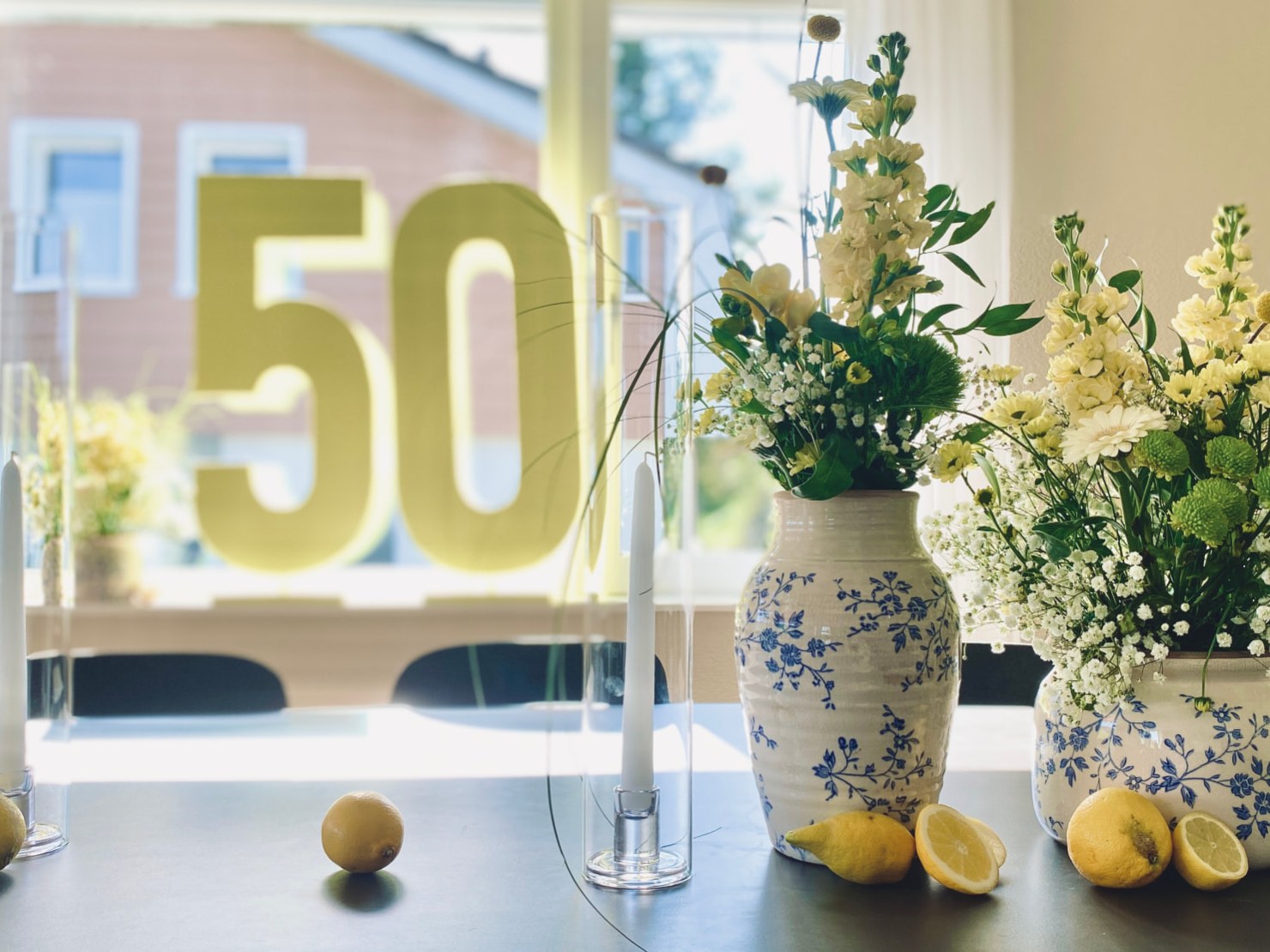 Gedeckter Tisch mit gelben Ziffern 50 vor dem Fenster, Kerzenständern, Zitronen und blau-weissen Blumenvasen