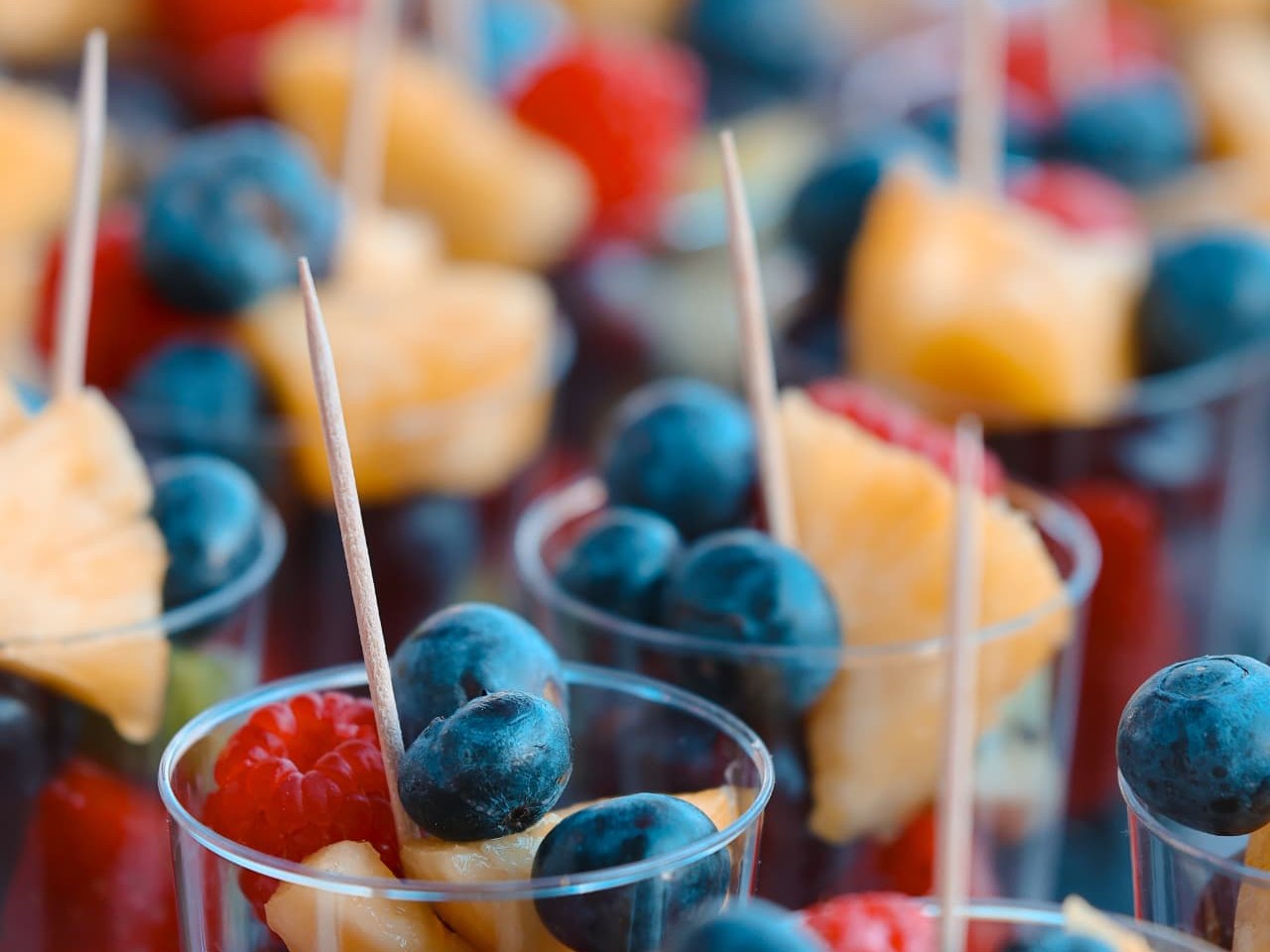 Fruchtbecher mit Blaubeeren, Himbeeren, Melonenstücken und Trauben, jeweils mit Holzspiessen