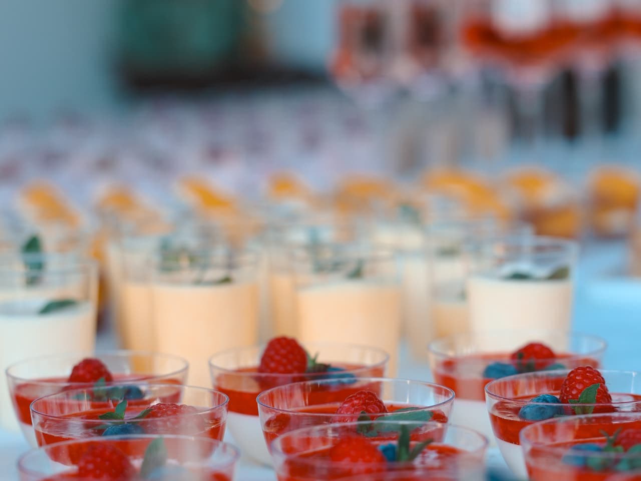 Dessertbecher mit Panna Cotta, rotem Fruchtspiegel, Himbeeren, Blaubeeren und Minze, dahinter hohe Gläser mit heller Creme