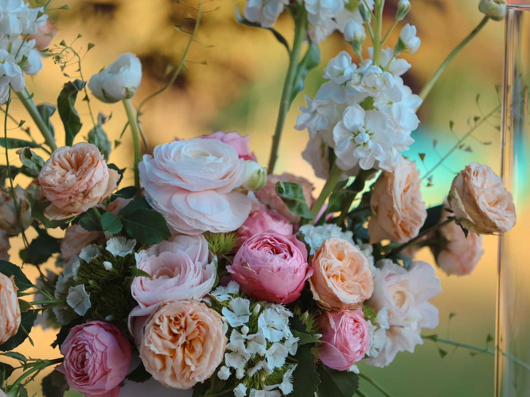 Nahaufnahme eines floralen Tischgestecks aus rosafarbenen und apricotfarbenen Rosen, Ranunkeln und weissen Blüten mit einer brennenden Kerze im Glaszylinder