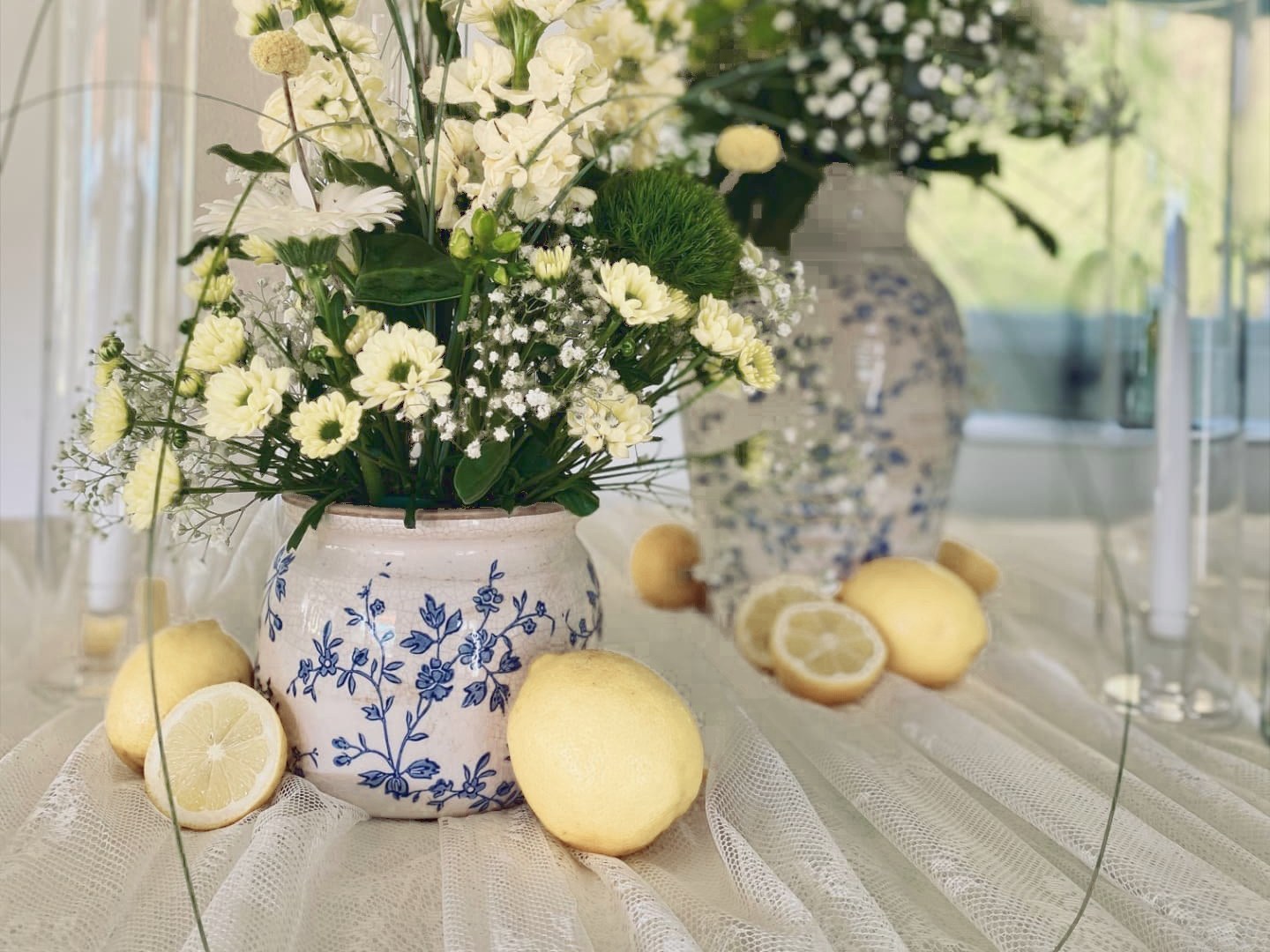 Gelb-weisse Blumen in blau-weissen Keramikvasen, mit Zitronen auf weissem Tüll dekoriert